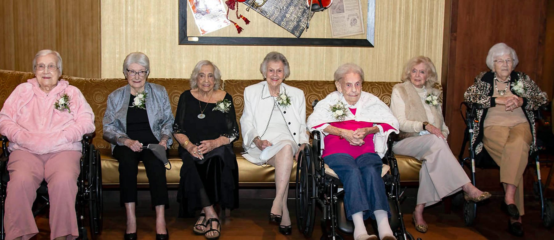 Seven elderly women, dressed nicely and wearing corsages, are seated together on a sofa and in wheelchairs, smiling at the camera in a warmly lit room with framed art on the wall behind them.