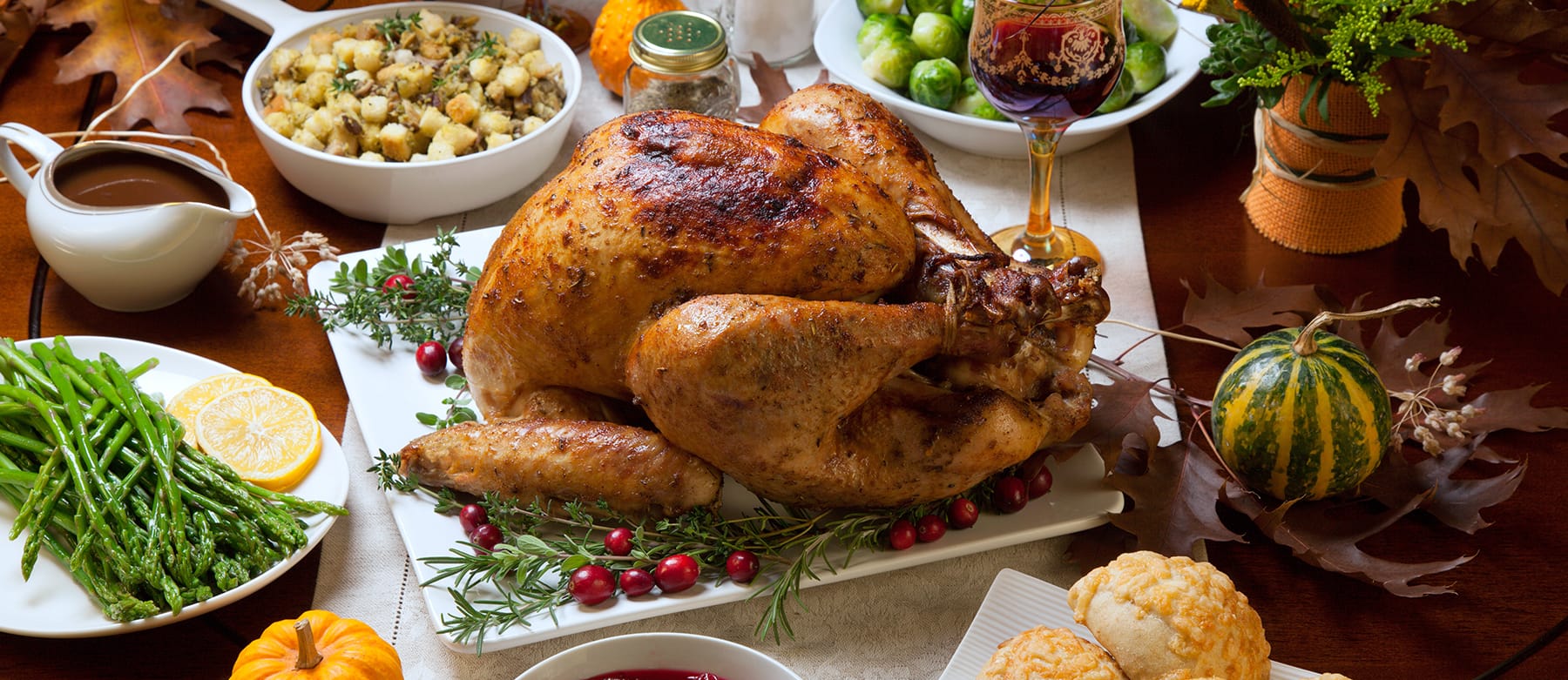 A roasted turkey garnished with herbs and cranberries sits on a platter, surrounded by side dishes like asparagus, Brussels sprouts, stuffing, biscuits, gravy, and drinks on a decorated autumn table.