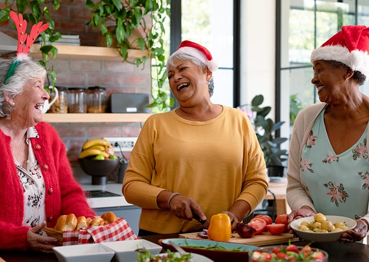 Three older women wearing festive hats laugh together while preparing food in a bright kitchen. One is slicing vegetables, and the counter is filled with bowls, bread, and holiday decorations.
