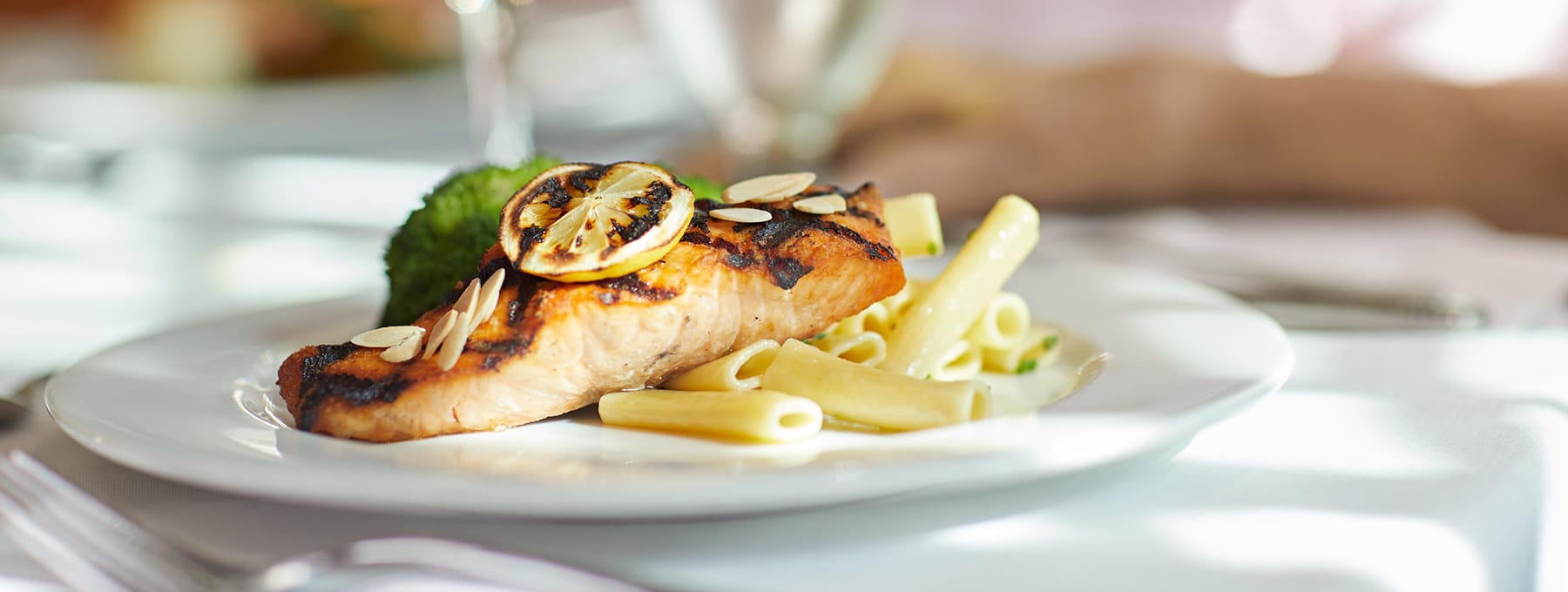 A plate of grilled salmon topped with lemon slices and almond slivers, served with penne pasta and a side of broccoli, set on a white tablecloth in a bright dining setting.