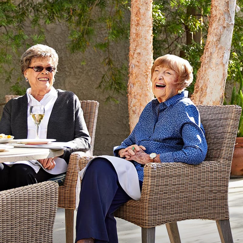 Two elderly women sit outside at a table, smiling and laughing. One has a glass of white wine; both wear glasses and light jackets. Trees and greenery are in the background, creating a cheerful, relaxed atmosphere.