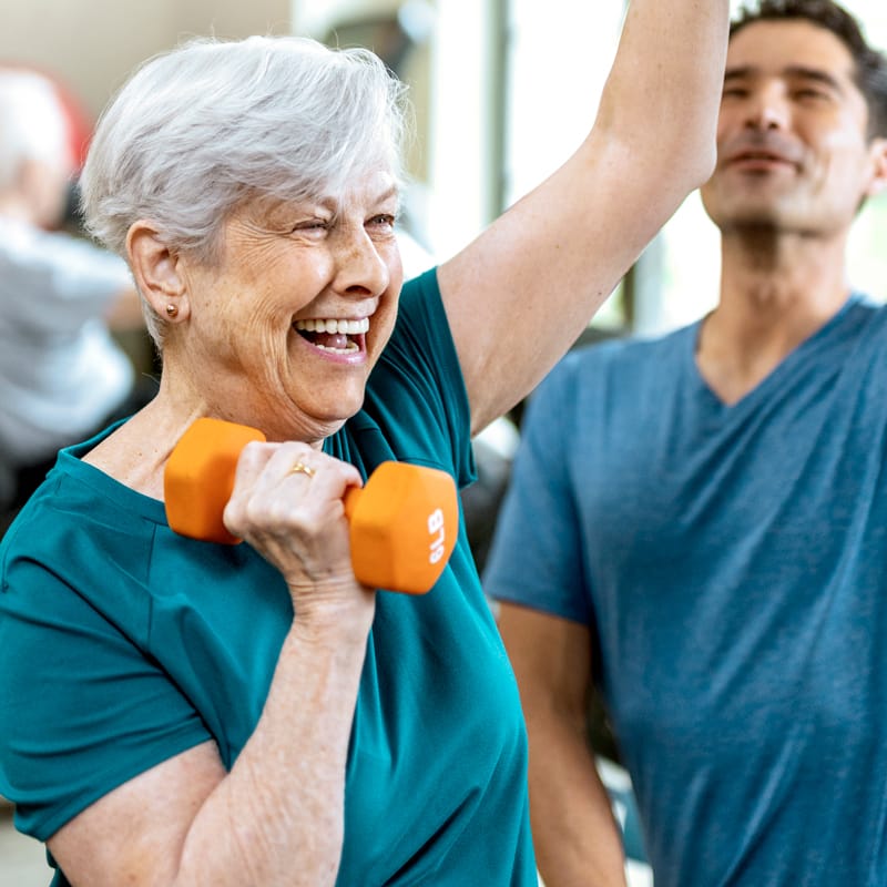 Smiling older woman with short gray hair lifts an orange dumbbell while exercising indoors, with a man in a blue shirt standing beside her in the background.