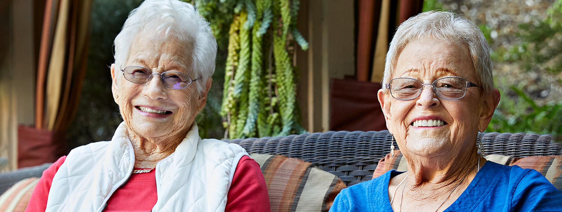 Two elderly women with short gray hair and glasses sit close together on an outdoor wicker sofa, smiling at the camera. They are surrounded by plants and appear to be enjoying a pleasant day.