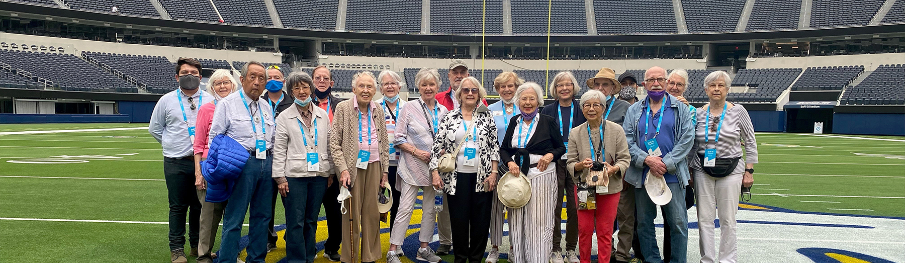 A group of older adults stands together on a football field in a large, mostly empty stadium, smiling for a photo. Most wear lanyards and casual clothing; some hold hats or cameras.