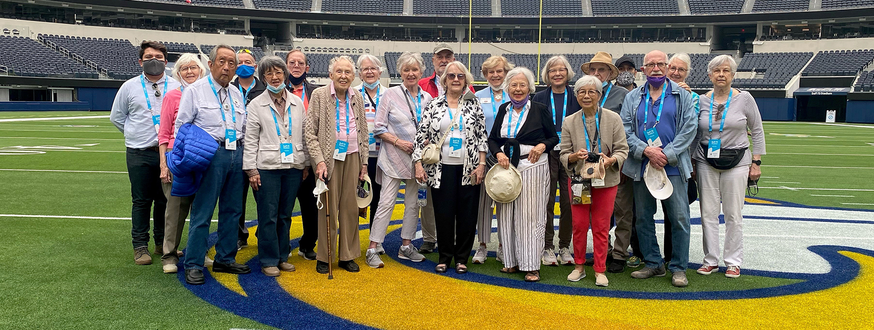 A group of about twenty older adults poses for a photo on a football field inside a large stadium, standing on the teams logo. Most wear name tags and casual clothing, and some hold hats or cameras.