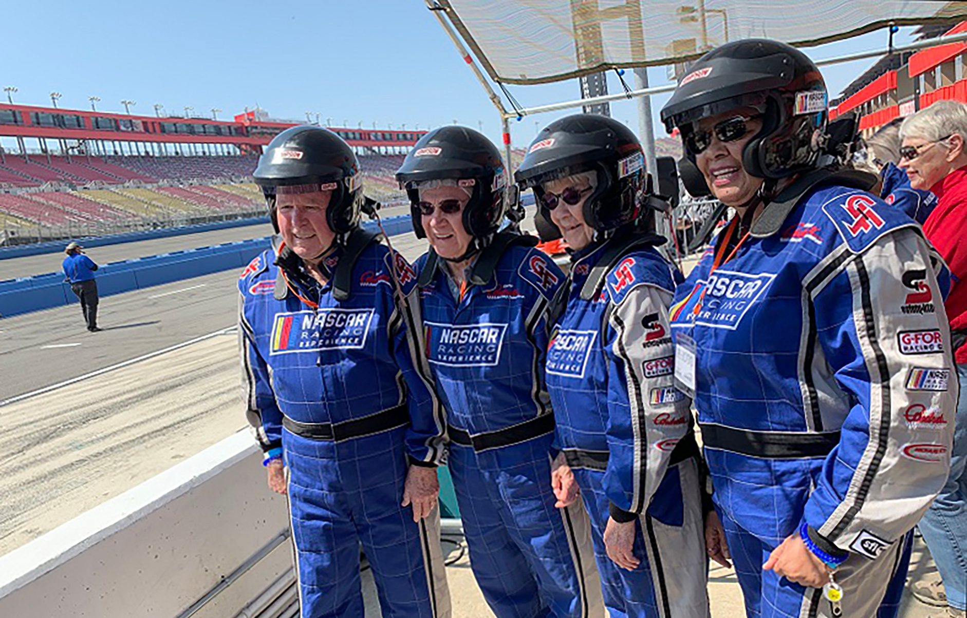 Four people in blue NASCAR racing suits and helmets stand side by side, their smiles reflecting creativity and teamwork at a racetrack with empty stands in the background on a sunny day.