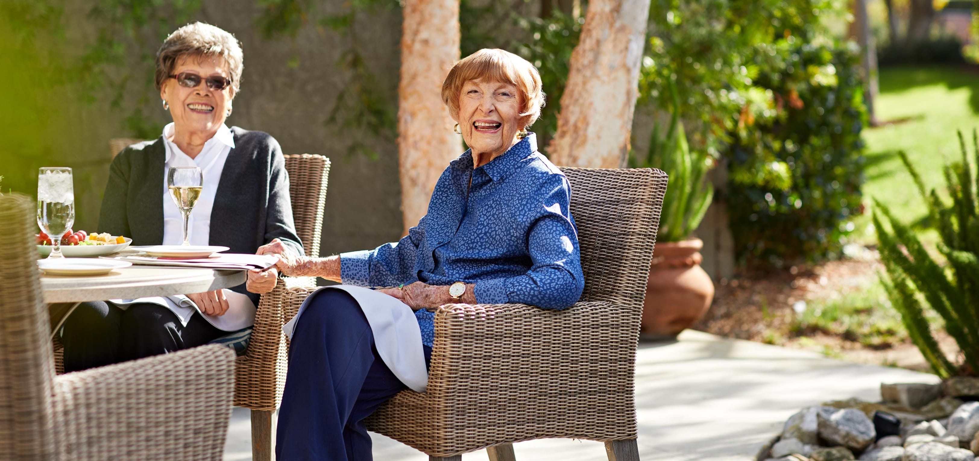 Two older women sit outside at a table, smiling and enjoying drinks and food on a sunny day. The scene is relaxed, with greenery and trees in the background.