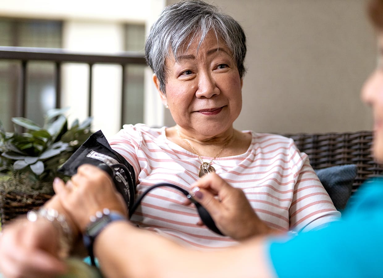 An older woman with short gray hair sits on a couch, smiling while another person checks her blood pressure with a cuff. They appear to be outdoors, and a plant is visible in the background.