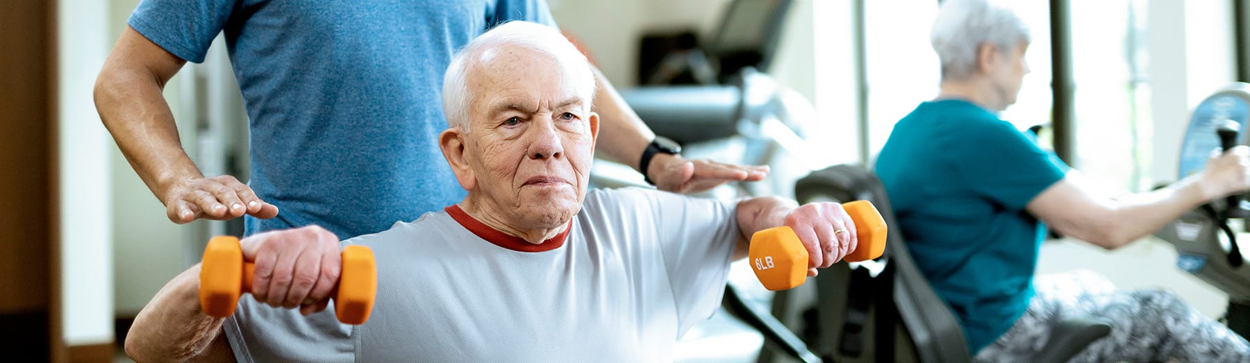 An older man lifts orange dumbbells while a trainer assists him. In the background, a woman exercises on a stationary bike. The setting appears to be a bright, modern gym or fitness center.