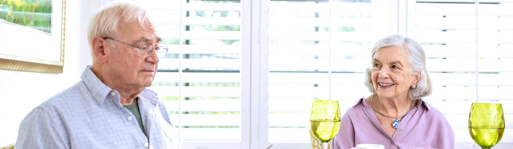 An elderly man and woman sit at a table with yellow-tinted glasses in front of them. The woman is smiling at the man, and they are in a bright room with white shutters on the windows.
