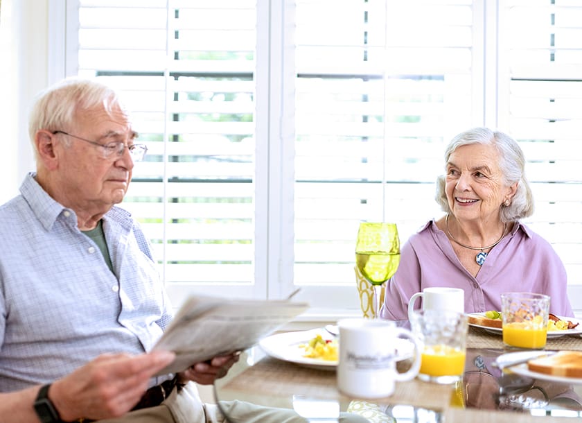 An elderly man reads a newspaper while seated at a breakfast table with an elderly woman who is smiling at him. The table has plates of food, glasses of orange juice, and mugs, with sunlight streaming through window blinds.