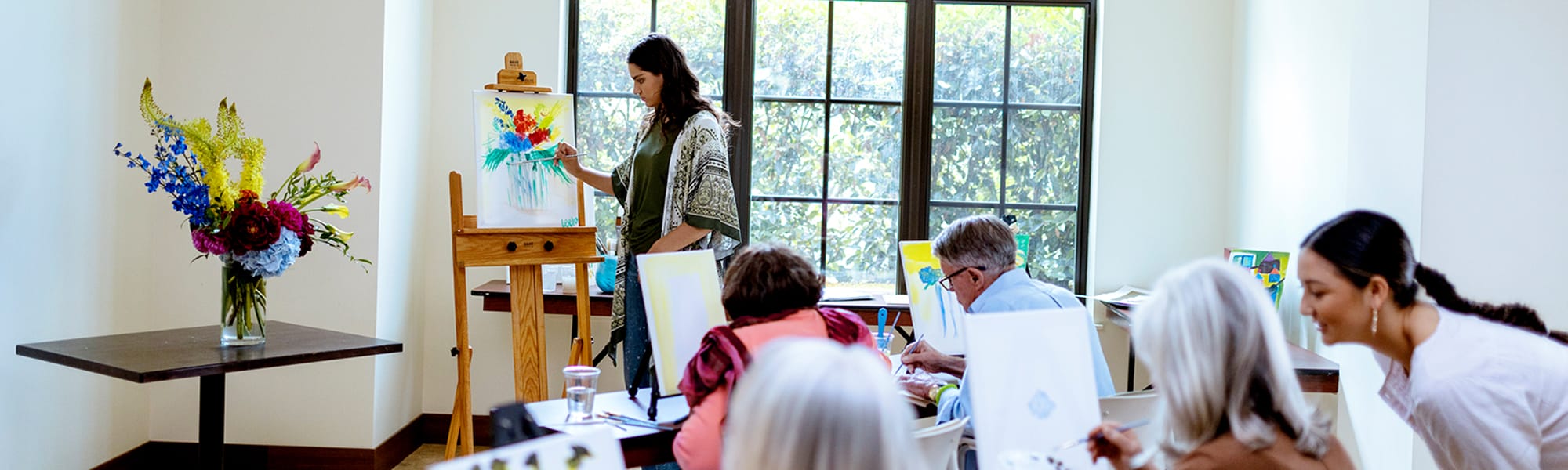A group of adults participates in a painting class in a bright room. One person stands at an easel painting a colorful floral piece, while others sit at tables working on their own artwork. A vase with flowers sits on a nearby table.