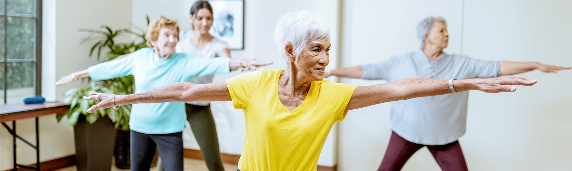 Four senior women in casual workout clothes are practicing yoga together indoors, standing in a row with arms outstretched in a warrior pose. They appear focused and engaged in the activity.