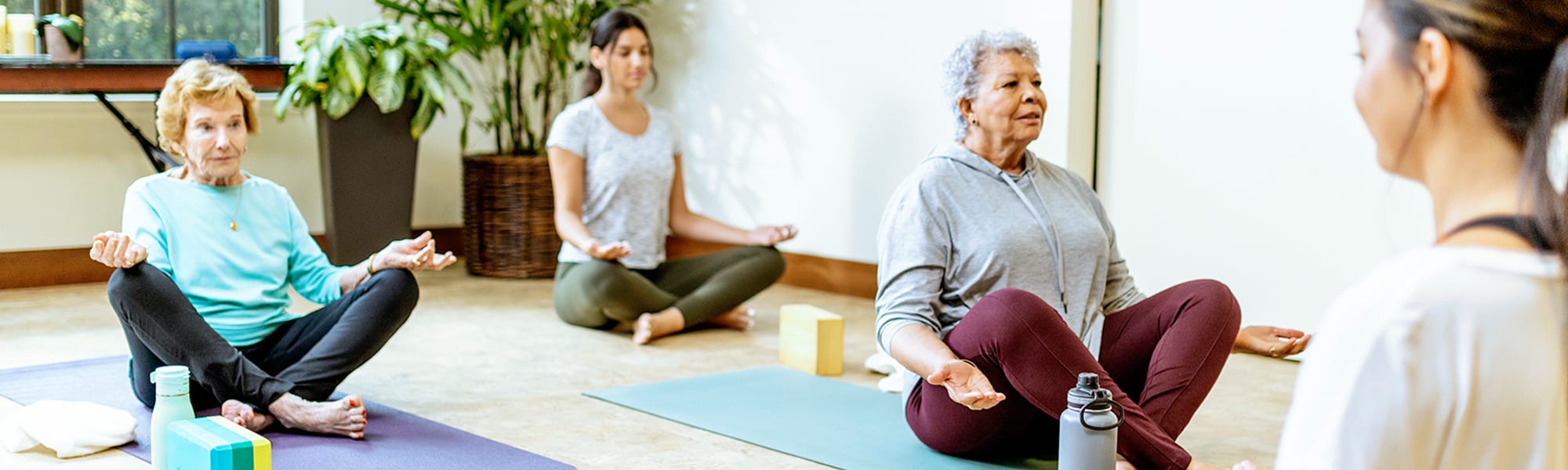 Three women sit cross-legged on yoga mats in a bright room, meditating with eyes closed and hands resting on knees. A fourth person, possibly an instructor, faces them. Yoga blocks and water bottles are nearby.