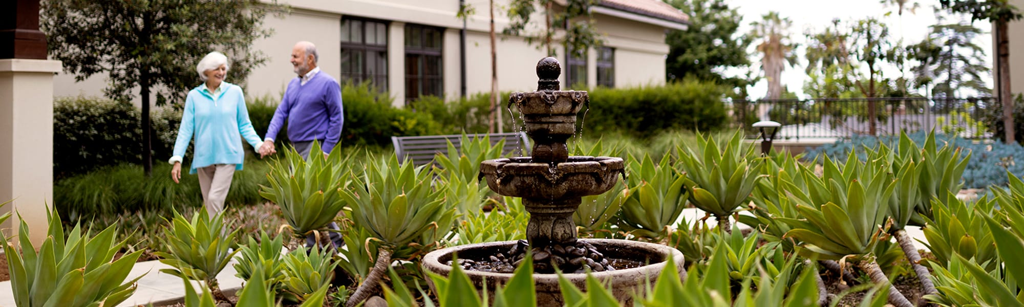 An elderly couple holding hands walks through a garden with green plants and a stone fountain in the foreground, near a building with large windows.