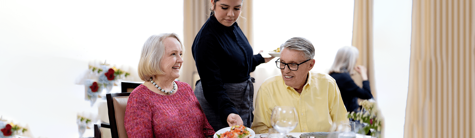 An older woman and man sit at a dining table smiling as a waitress serves them a plate of food, showcasing creativity in presentation. The room is bright with large windows and vases of flowers in the background.