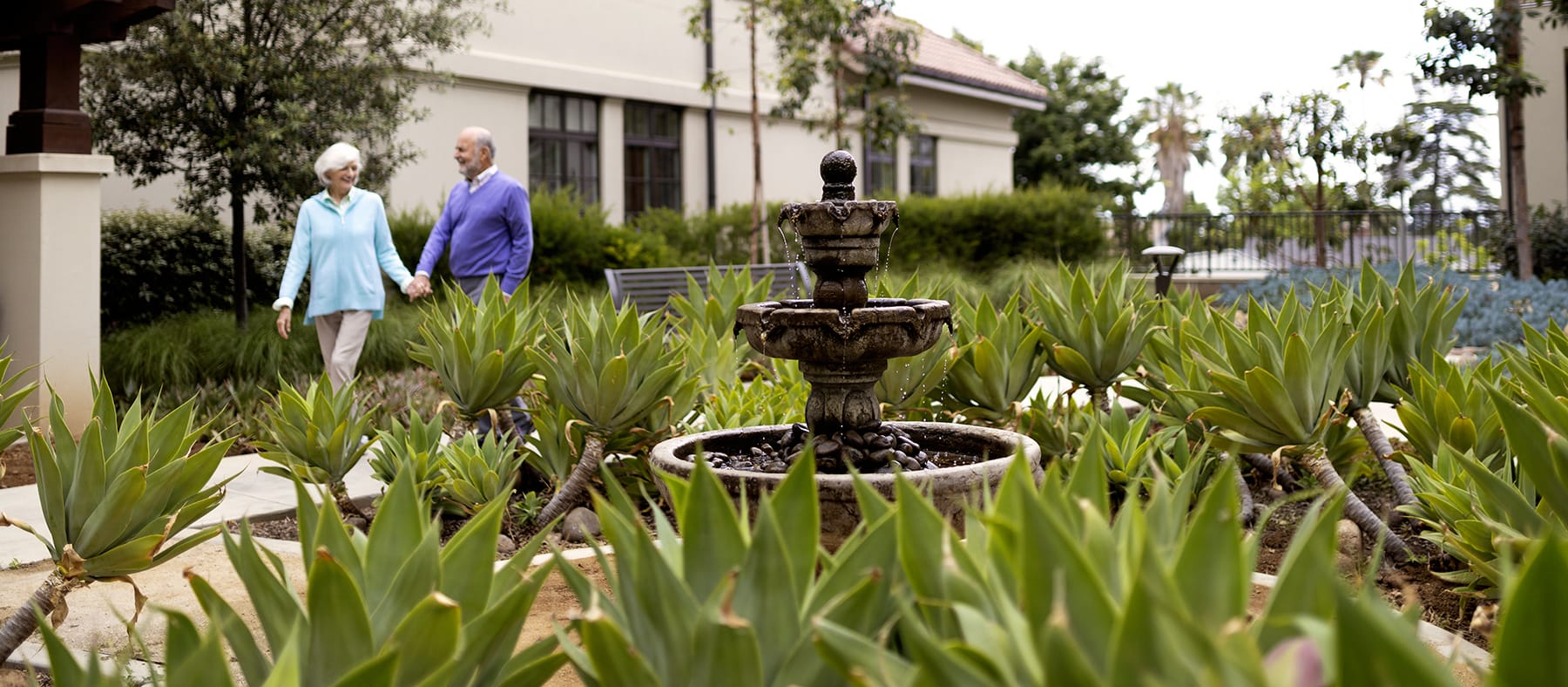 An elderly couple holds hands while walking through a garden with lush green plants and a stone fountain, next to a building with large windows.