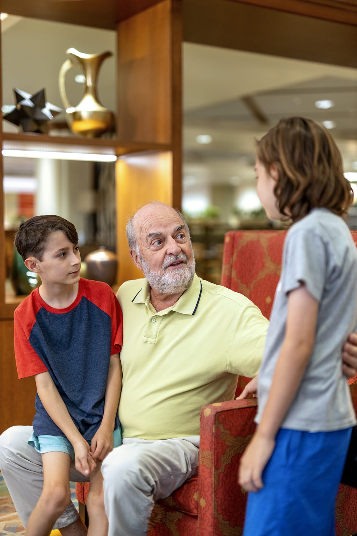 An older man with a white beard sits in a red chair, talking to two young boys. One boy sits on his knee while the other stands in front of them. They are indoors, and the background shows shelves and decorative items.