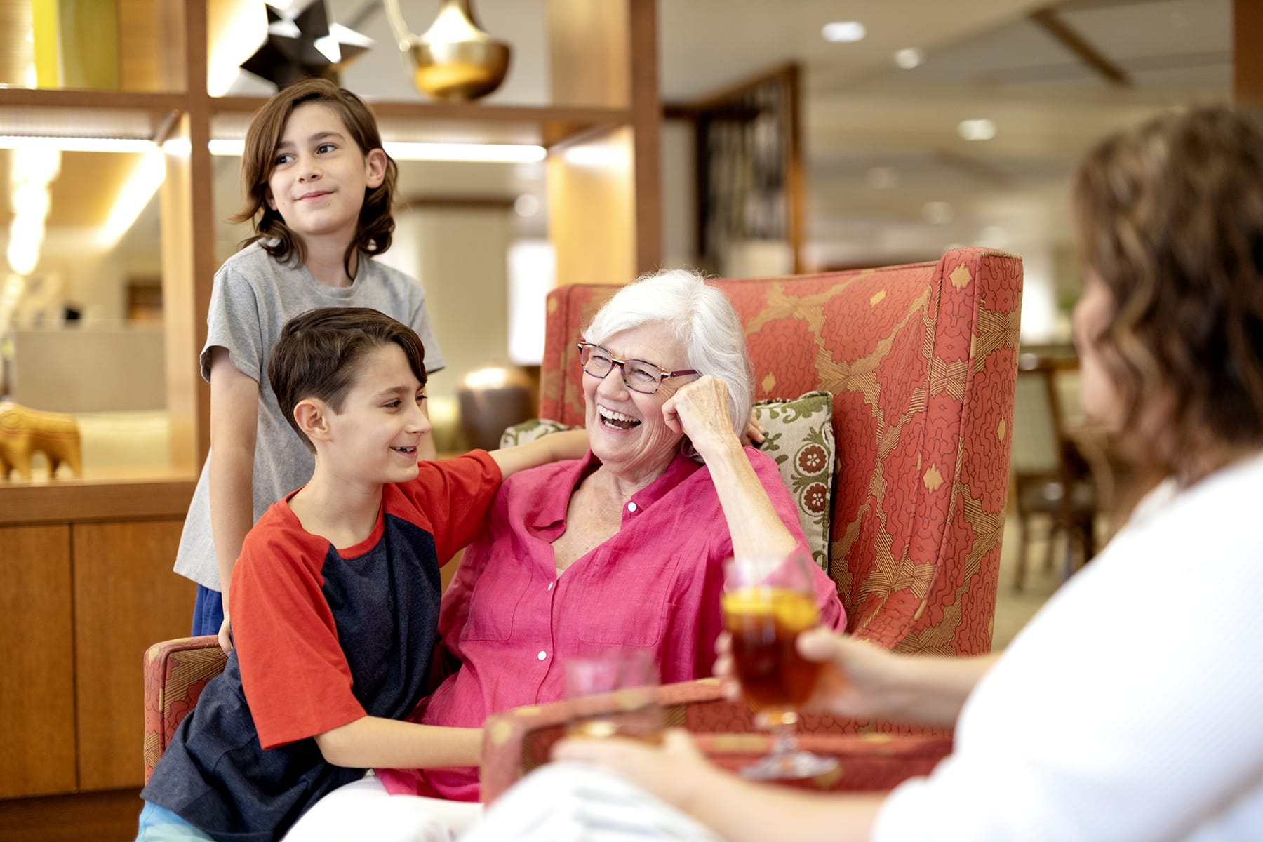 An elderly woman in a pink shirt smiles and laughs while sitting in an armchair, surrounded by two children and an adult holding a drink. The group appears happy in a cozy, well-lit indoor setting.