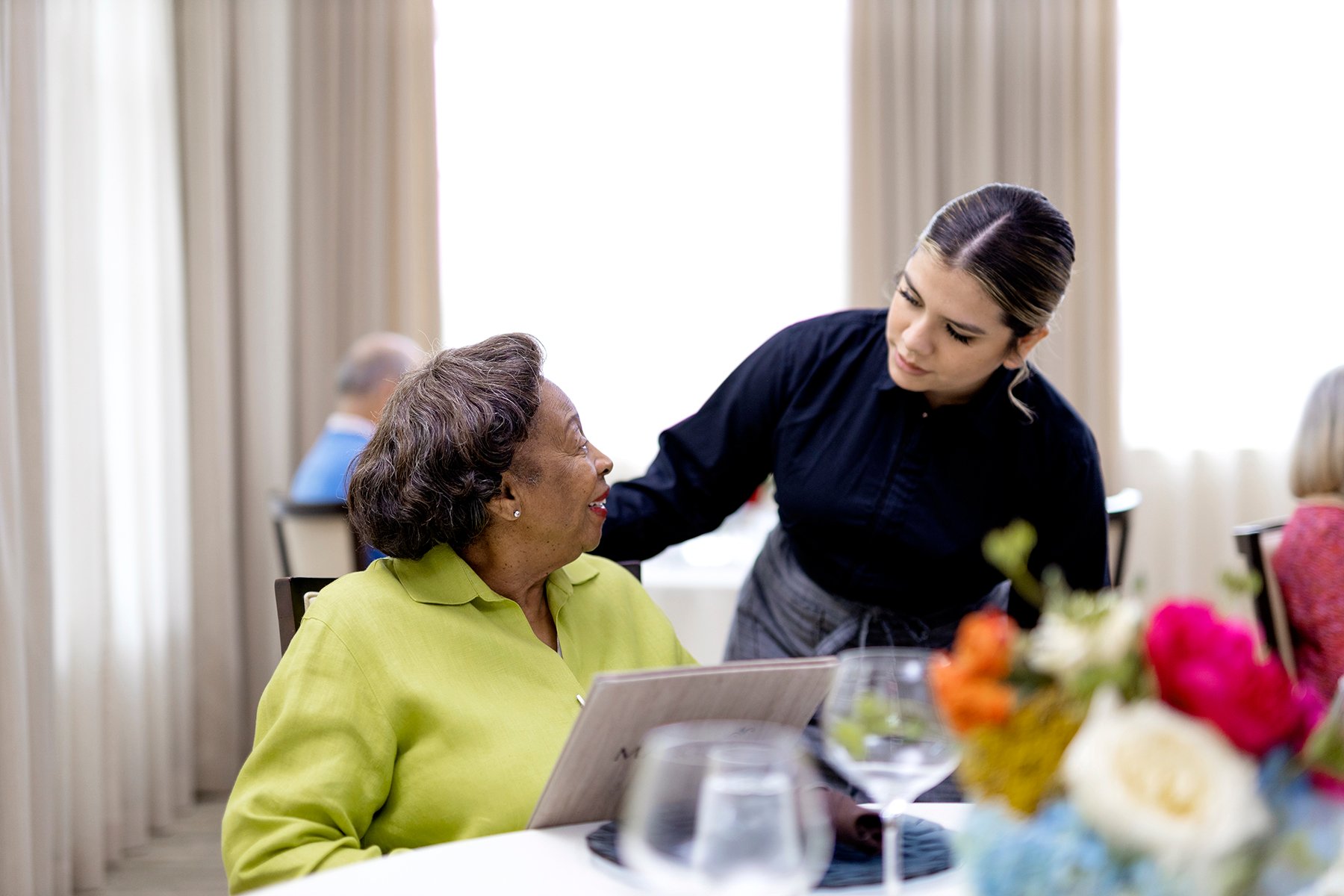 An elderly woman in a green blouse sits at a table with a menu, smiling and talking to a young waitress standing beside her. Bright flowers and glassware are on the table, with soft light coming through curtains in the background.