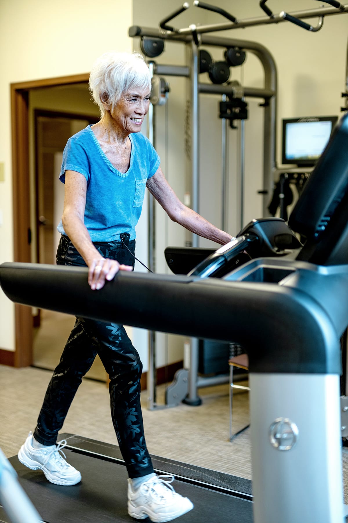 An older woman with short white hair, wearing a blue t-shirt, patterned pants, and white sneakers, walks on a treadmill in a gym, smiling and appearing active and healthy.