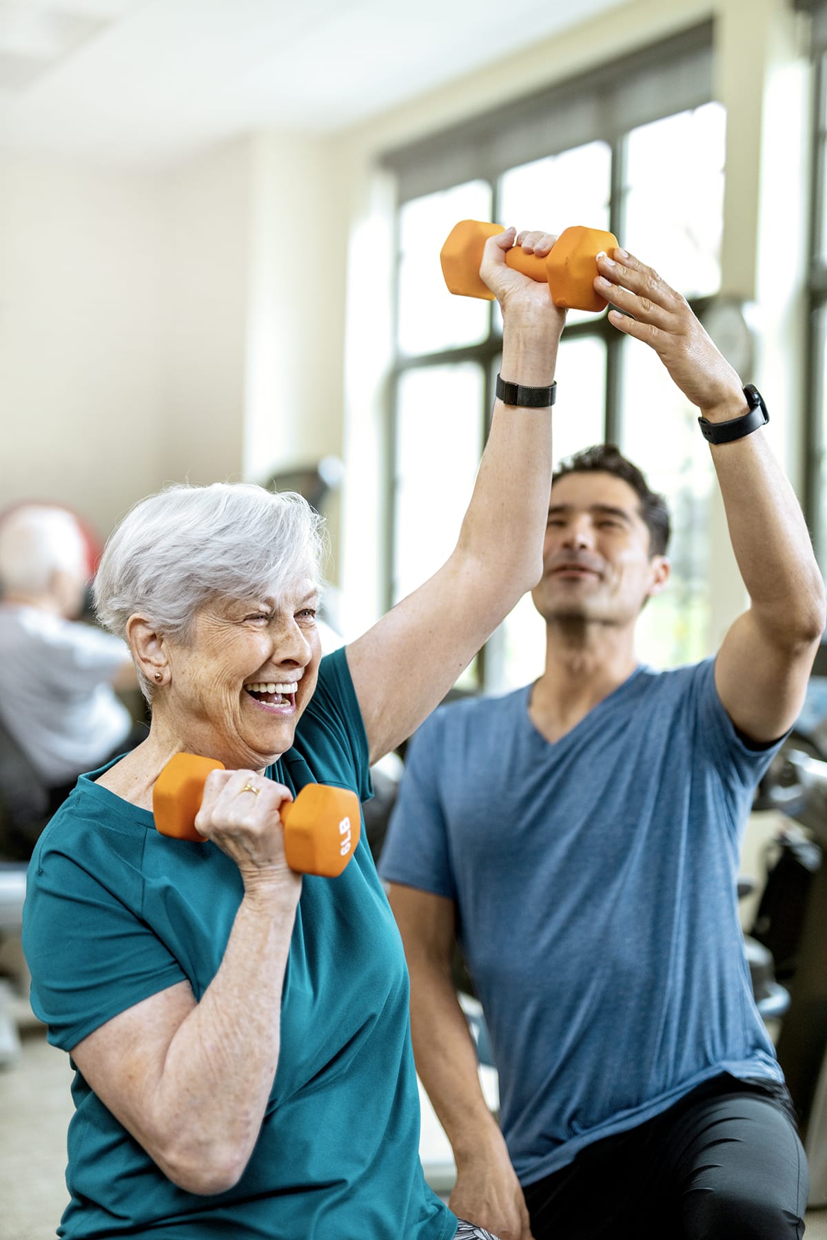 An older woman with short gray hair lifts orange dumbbells while smiling, assisted by a younger man in a blue shirt at a bright indoor gym.