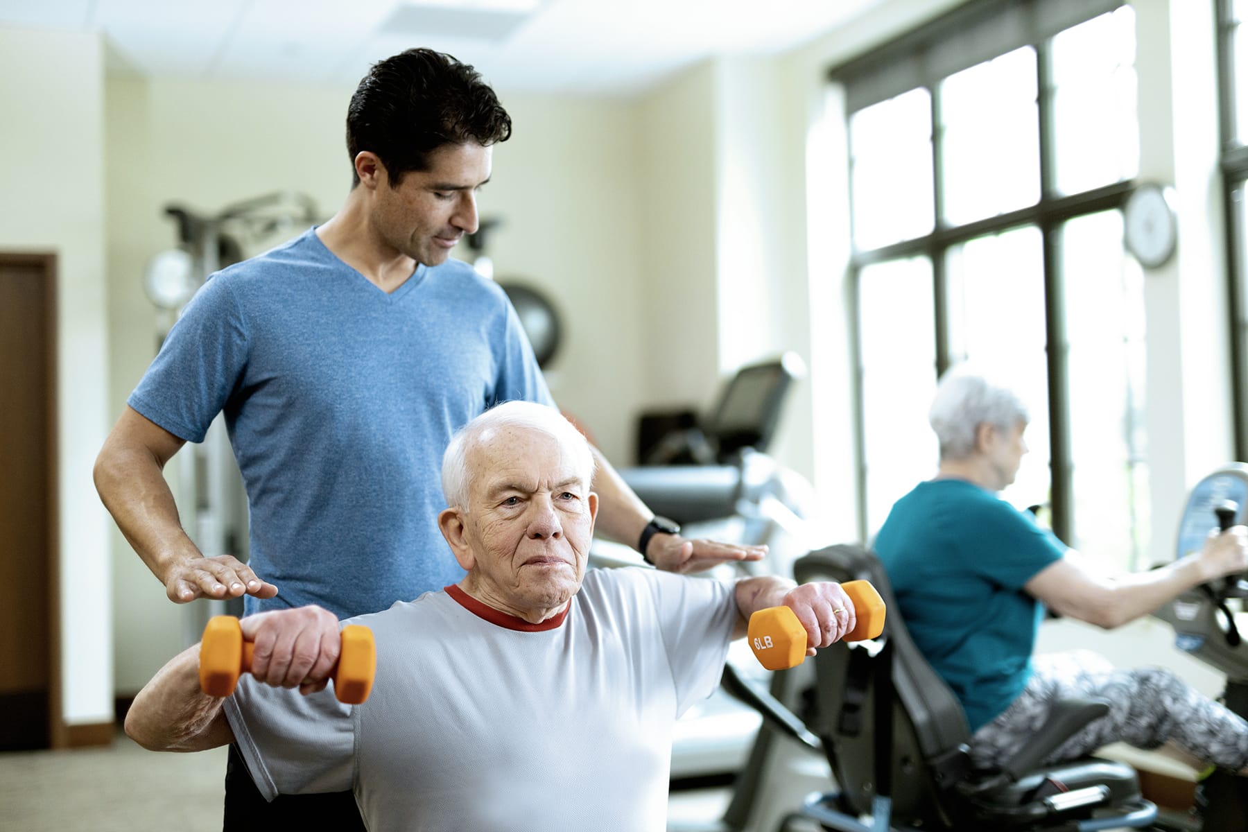 A young male trainer assists an older man lifting orange dumbbells indoors, while an elderly woman exercises on a stationary bike in the background.