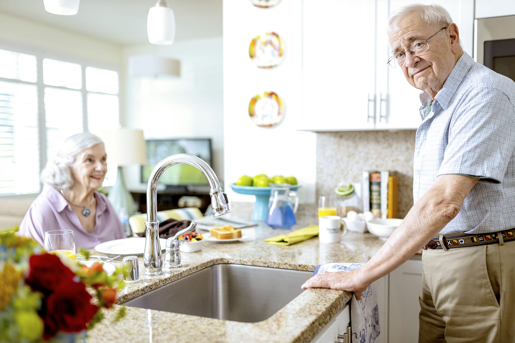 An elderly man stands at a kitchen counter, looking at the camera, while an elderly woman sits at a table in the background, smiling. The kitchen is bright and modern with various items neatly arranged.