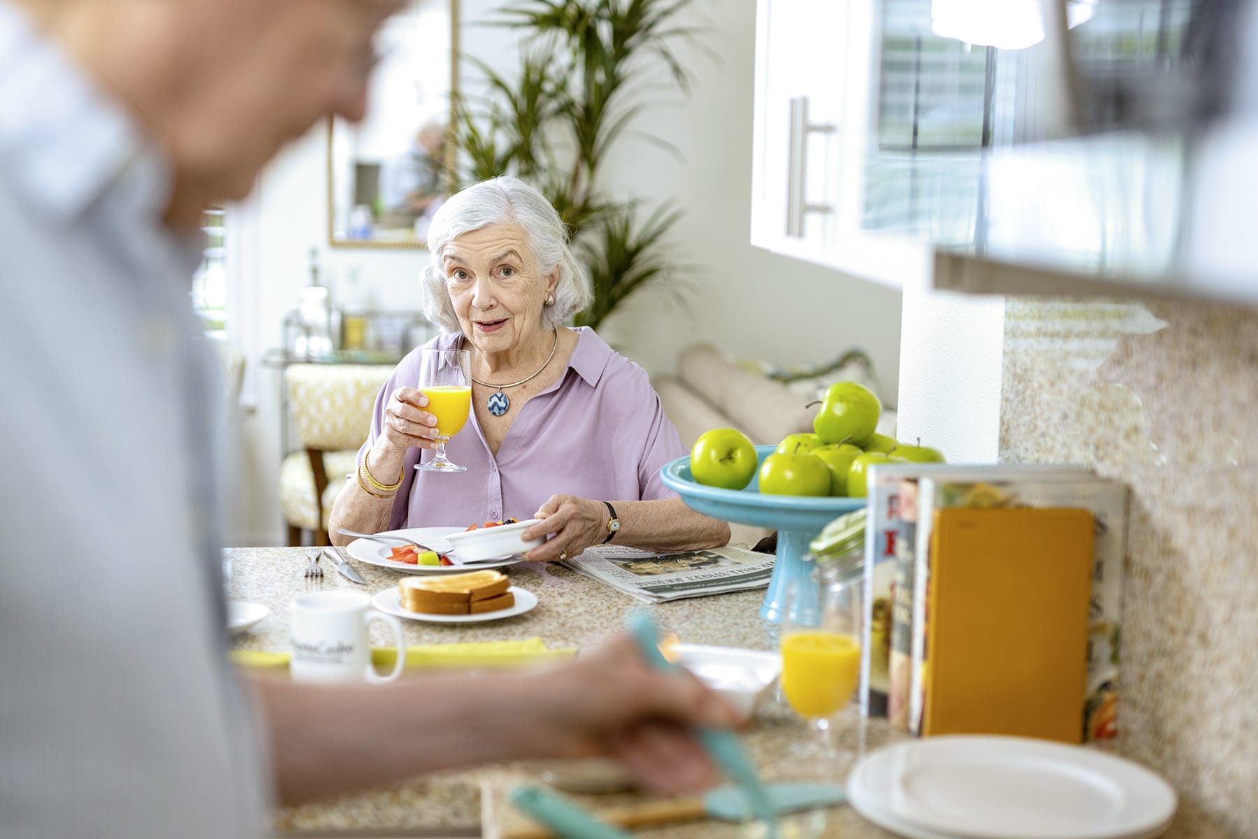 An older woman sits at a kitchen table smiling and holding a glass of orange juice, while an older man prepares food in the foreground. A bowl of green apples and breakfast items are on the counter.
