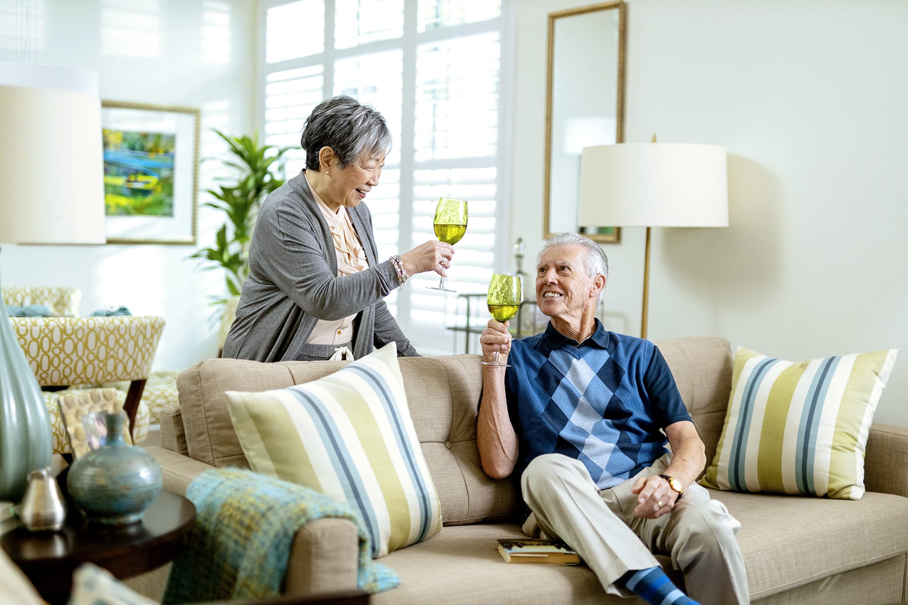 An older woman and man smile and clink green wine glasses while relaxing on a beige couch in a bright, modern living room with striped pillows and cheerful decor.