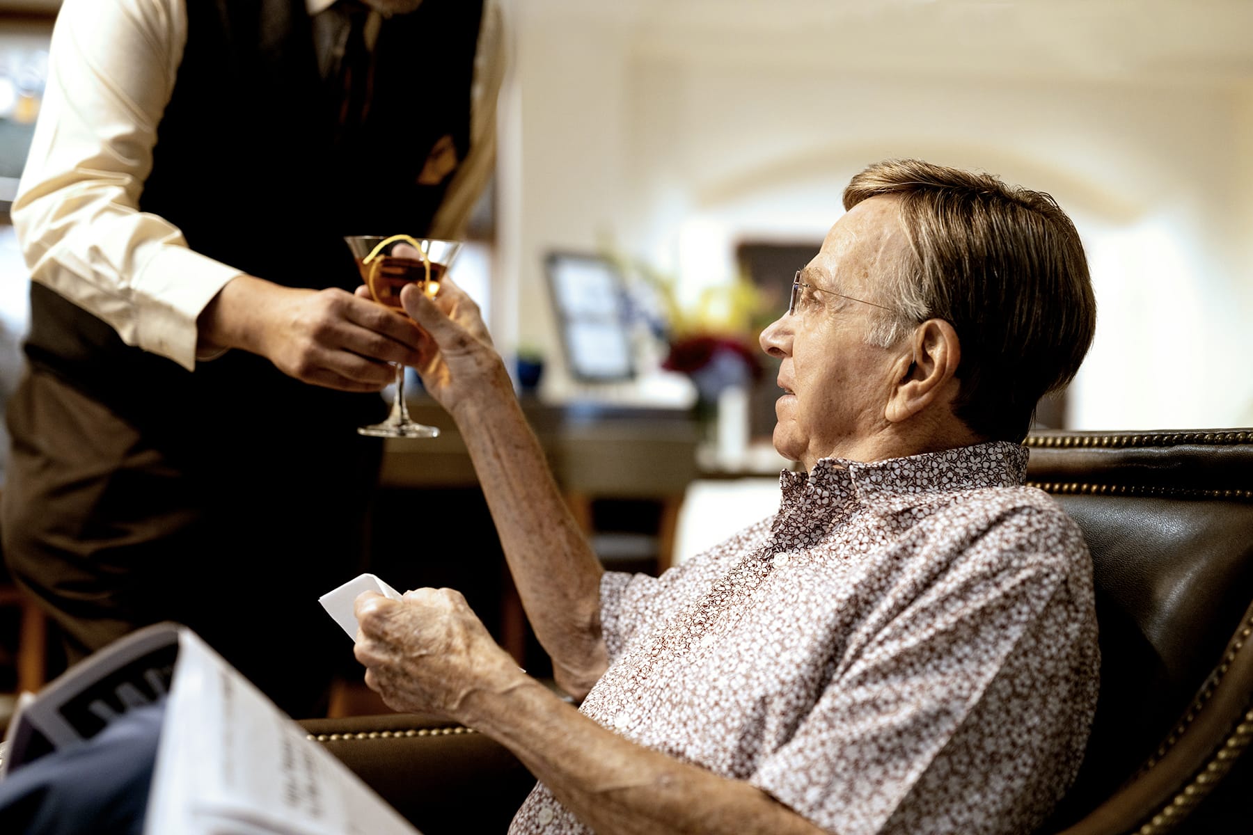An elderly man sitting in a chair receives a cocktail from a server. The man holds a card in one hand and looks up at the server, who wears a vest and white shirt. The setting appears to be a lounge or restaurant.
