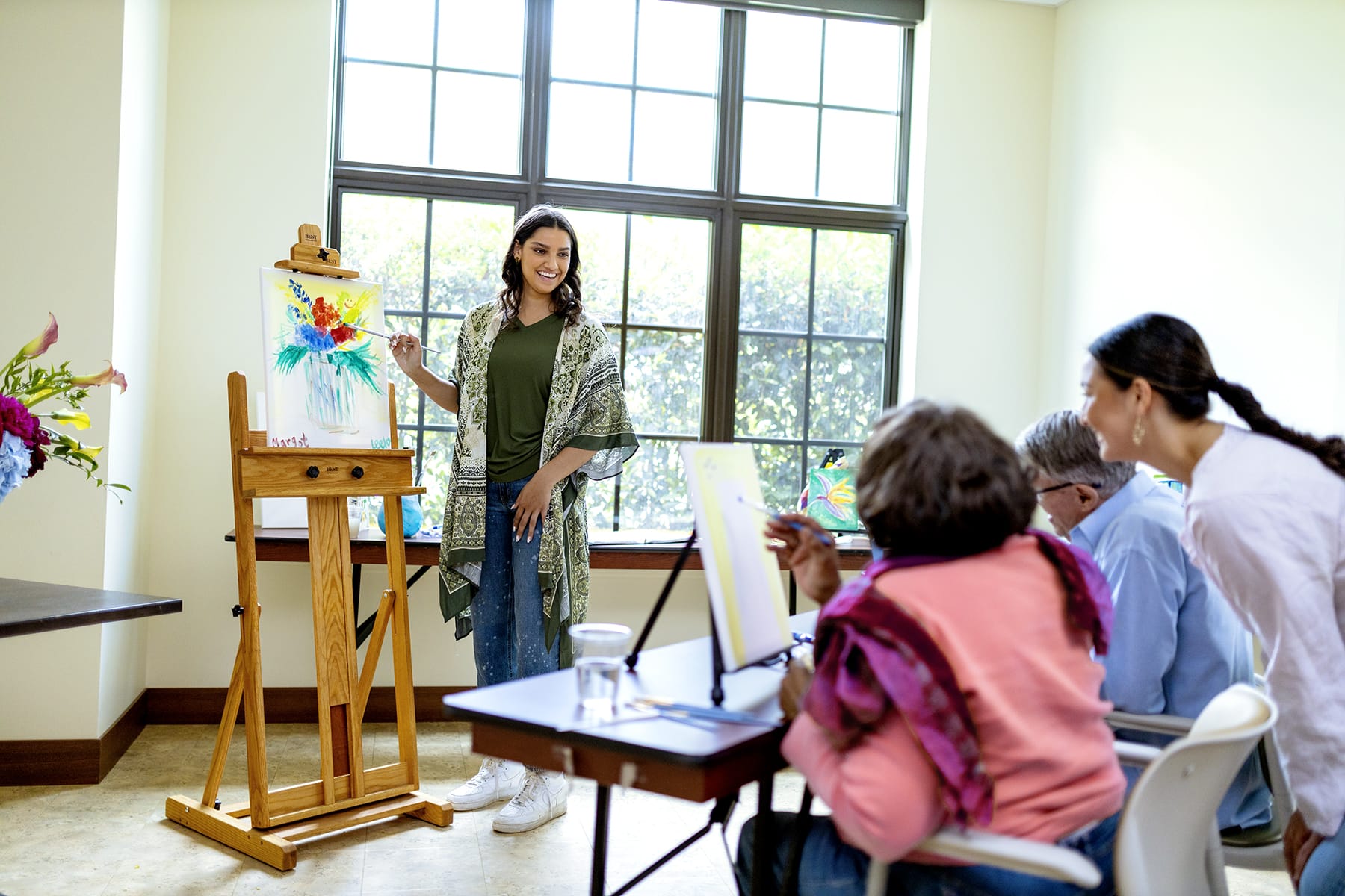 A woman stands by an easel displaying a colorful painting, smiling and teaching as three seated people watch and paint at a table in a bright room with large windows.