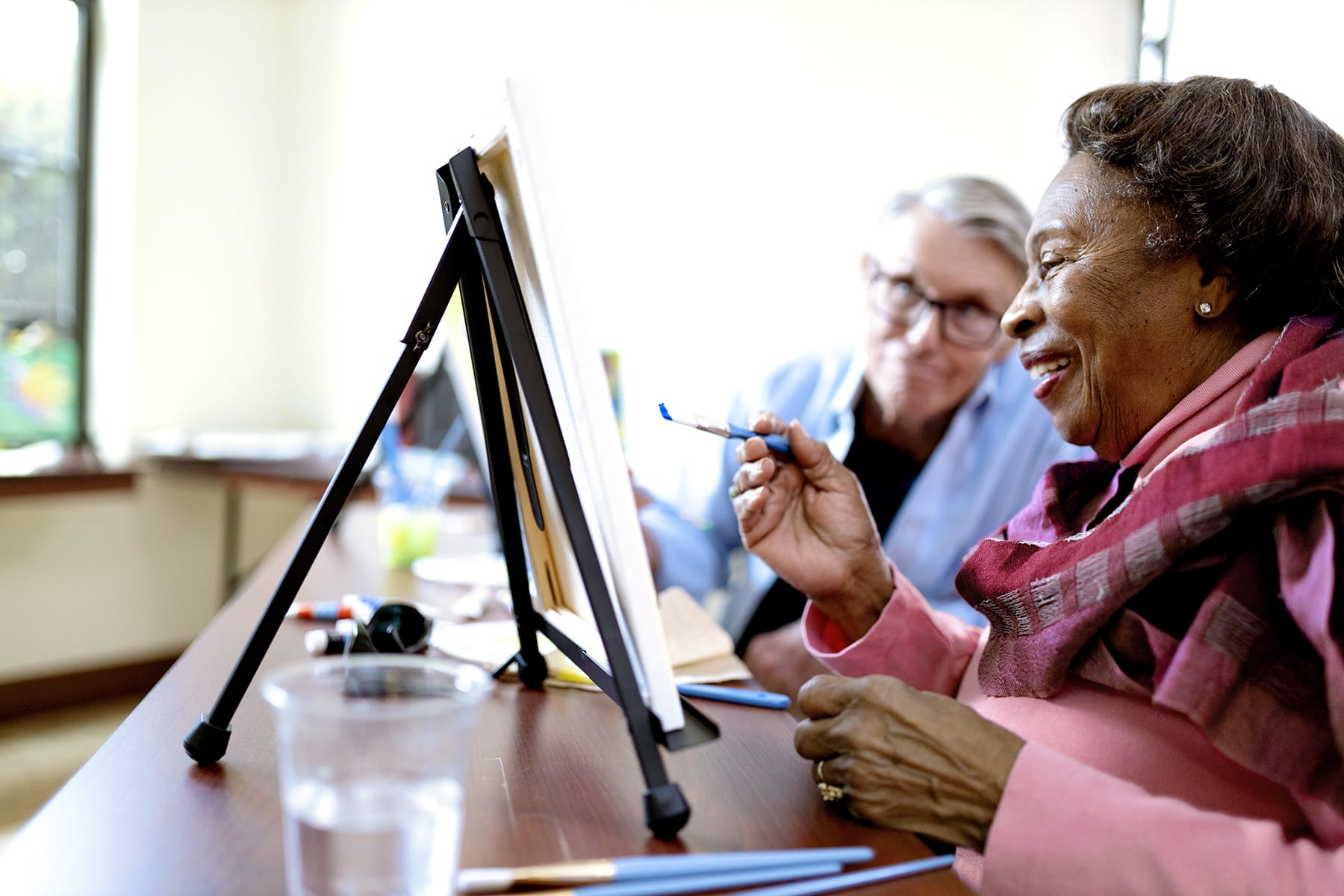 An elderly woman smiles while painting on a canvas at a table, with another person beside her offering support. Paintbrushes, a cup of water, and art supplies are visible on the table.