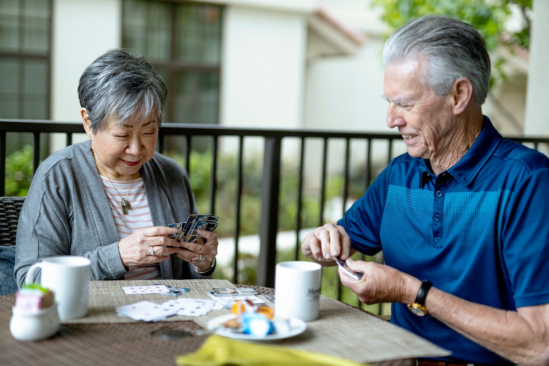 An older woman and man sit at an outdoor table, smiling as they play a card game. Two mugs and playing cards are on the table. They appear relaxed and happy, enjoying each others company.