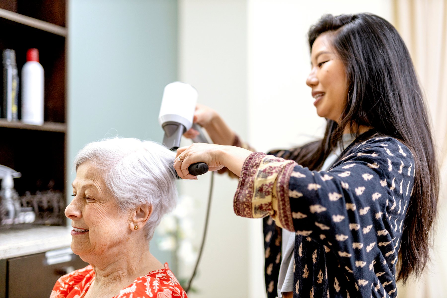 A young hairstylist blow-dries the short, white hair of an older woman who is smiling and wearing a red patterned blouse in a salon. Shelves with hair products are visible in the background.