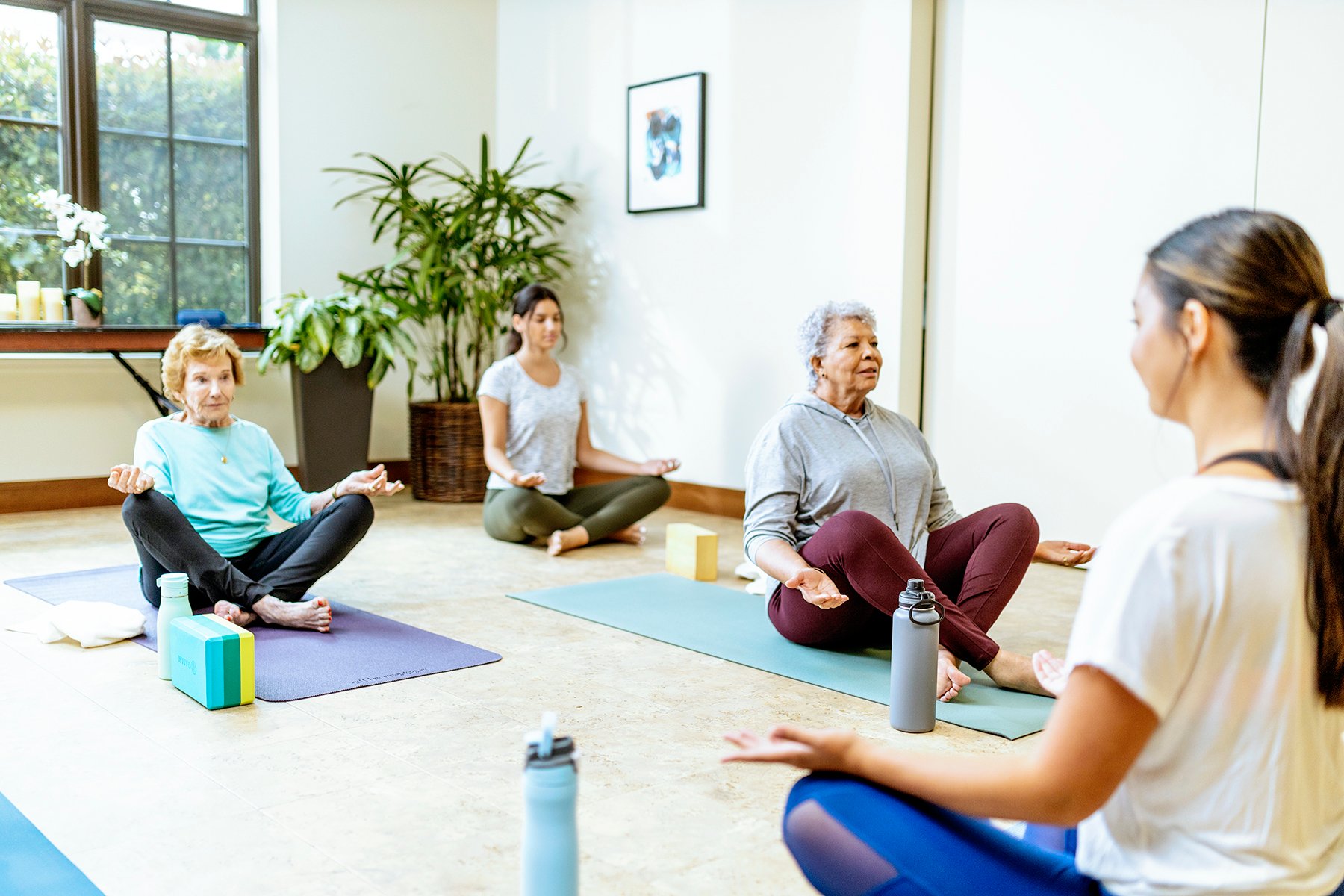 Three women of varying ages sit cross-legged on yoga mats during a yoga class, following an instructor at the front. The room is bright with natural light, potted plants, and exercise props nearby.