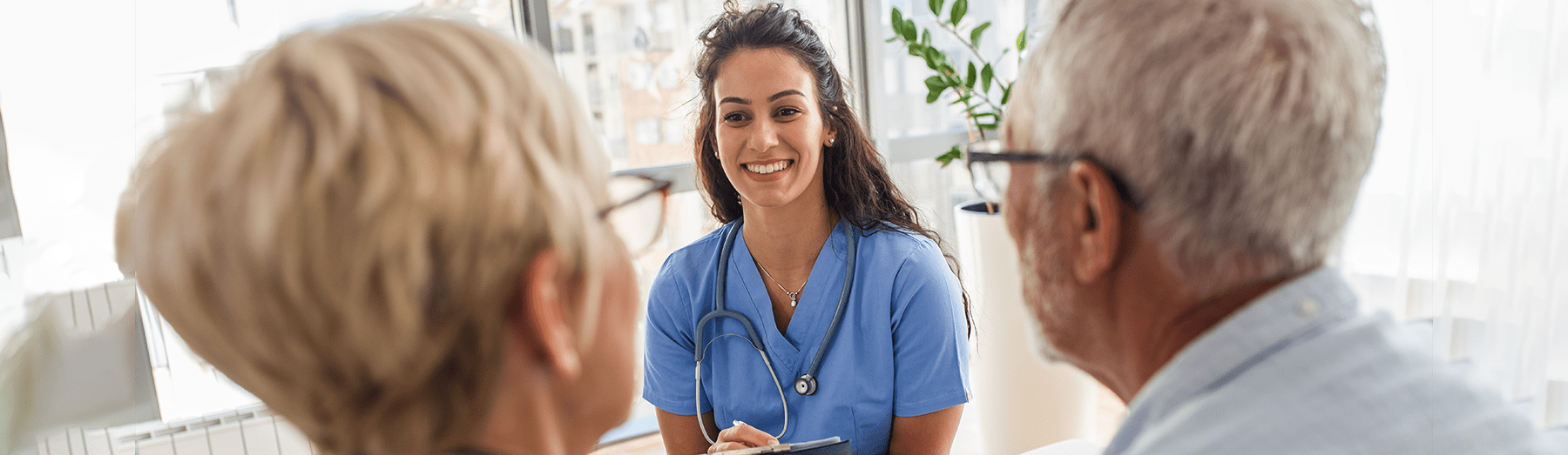 A smiling healthcare professional in blue scrubs sits with a stethoscope around her neck, talking to an older woman and man in a bright, sunlit room.