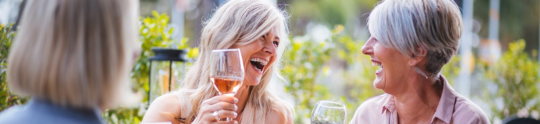 Three women sit outdoors, laughing and enjoying drinks together. The atmosphere is bright and cheerful, with greenery in the background. One woman holds a glass of rosé, and another woman has a glass of white wine.