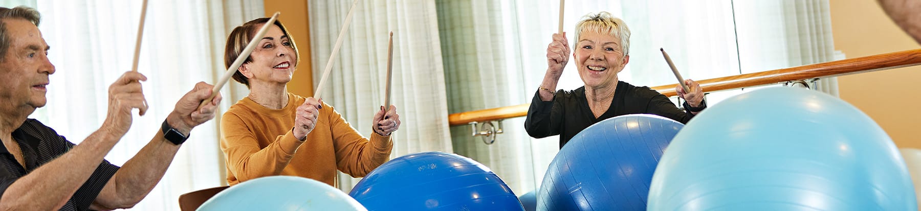 Three older adults smiling and exercising indoors, using drumsticks to hit large blue exercise balls placed in front of them. Light streams in through sheer curtains in the background.