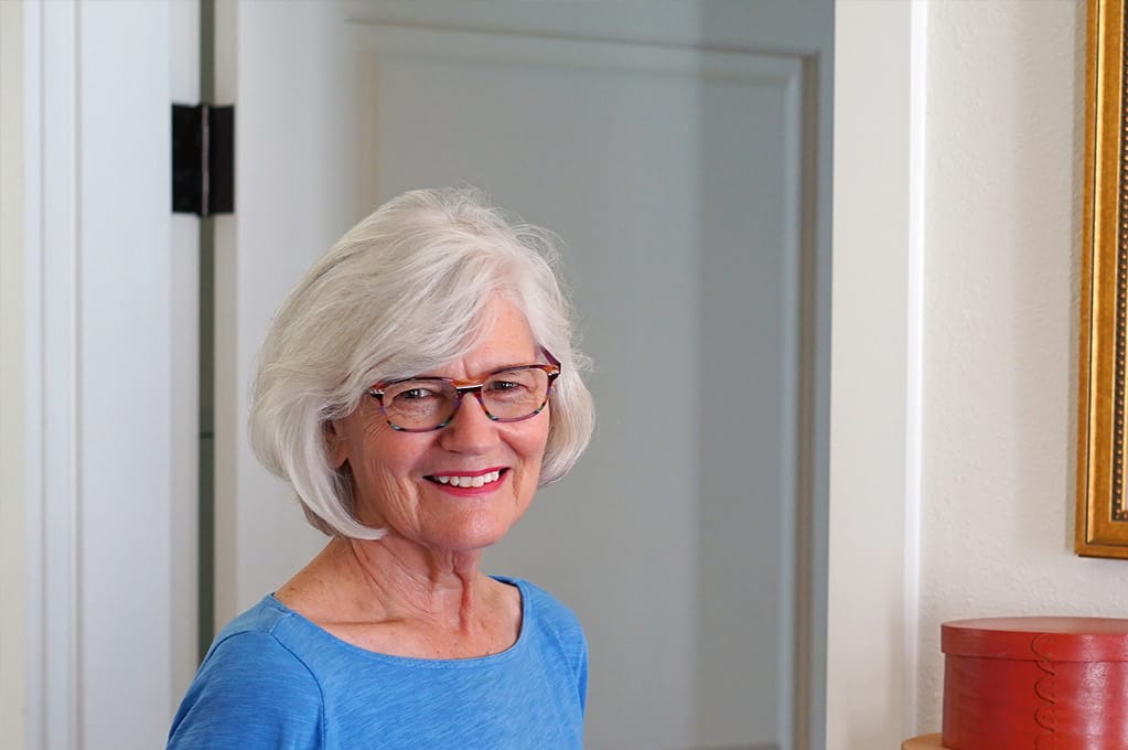 Smiling older woman with white hair and glasses, wearing a blue shirt, stands indoors near a doorway, where hints of creativity appear in the framed picture and red container partially visible nearby.