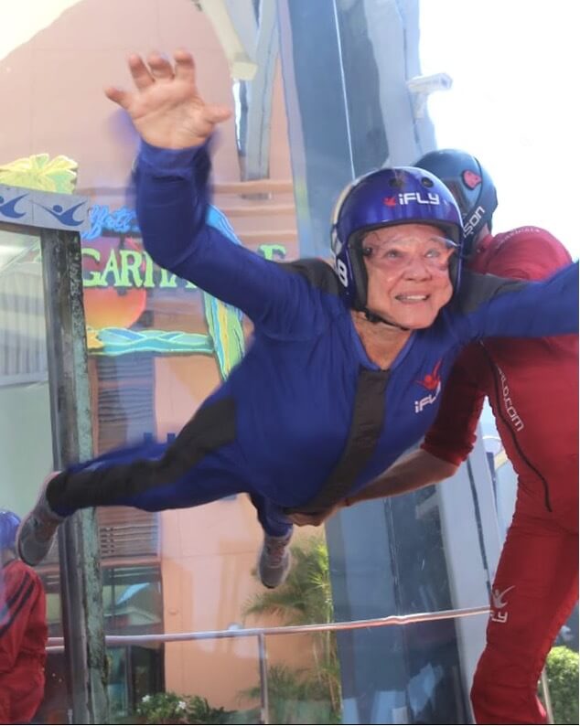 An older woman in a blue jumpsuit and helmet is indoor skydiving, supported by an instructor in red gear. She is smiling and has her arms extended, floating inside a vertical wind tunnel.