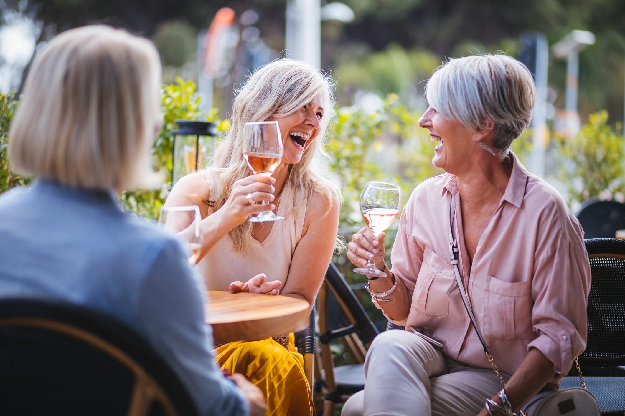 Three women with light hair sit outdoors at a table, smiling and laughing while holding glasses of rosé wine. The setting is bright and surrounded by greenery, creating a cheerful, relaxed atmosphere.