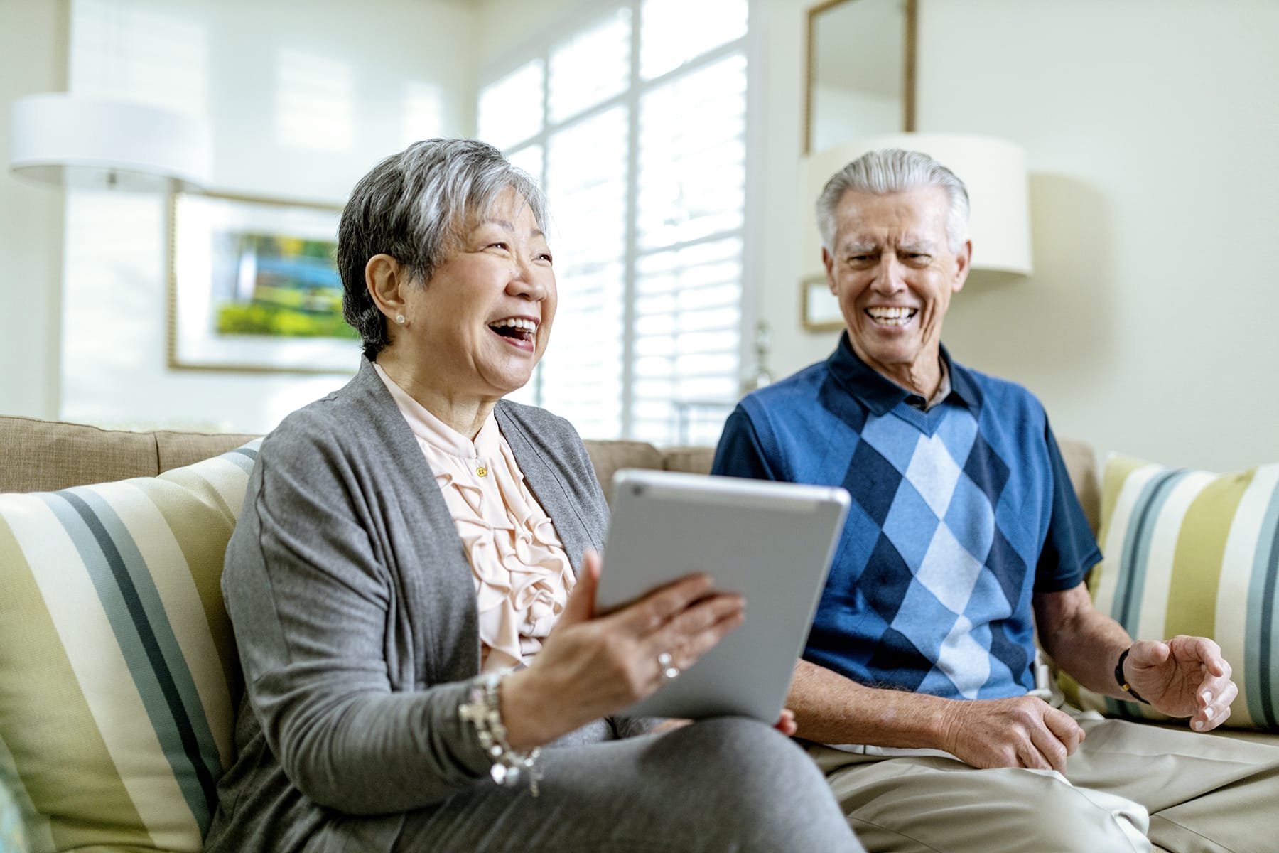 An elderly woman and man sit together on a couch, smiling and laughing while the woman holds a tablet. The bright room has striped pillows and sunlight streams through the windows behind them.