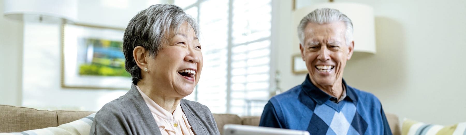 An older woman and man sit together on a couch, smiling and laughing while looking toward a tablet. The room is bright with natural light and decorated with a lamp and framed picture in the background.