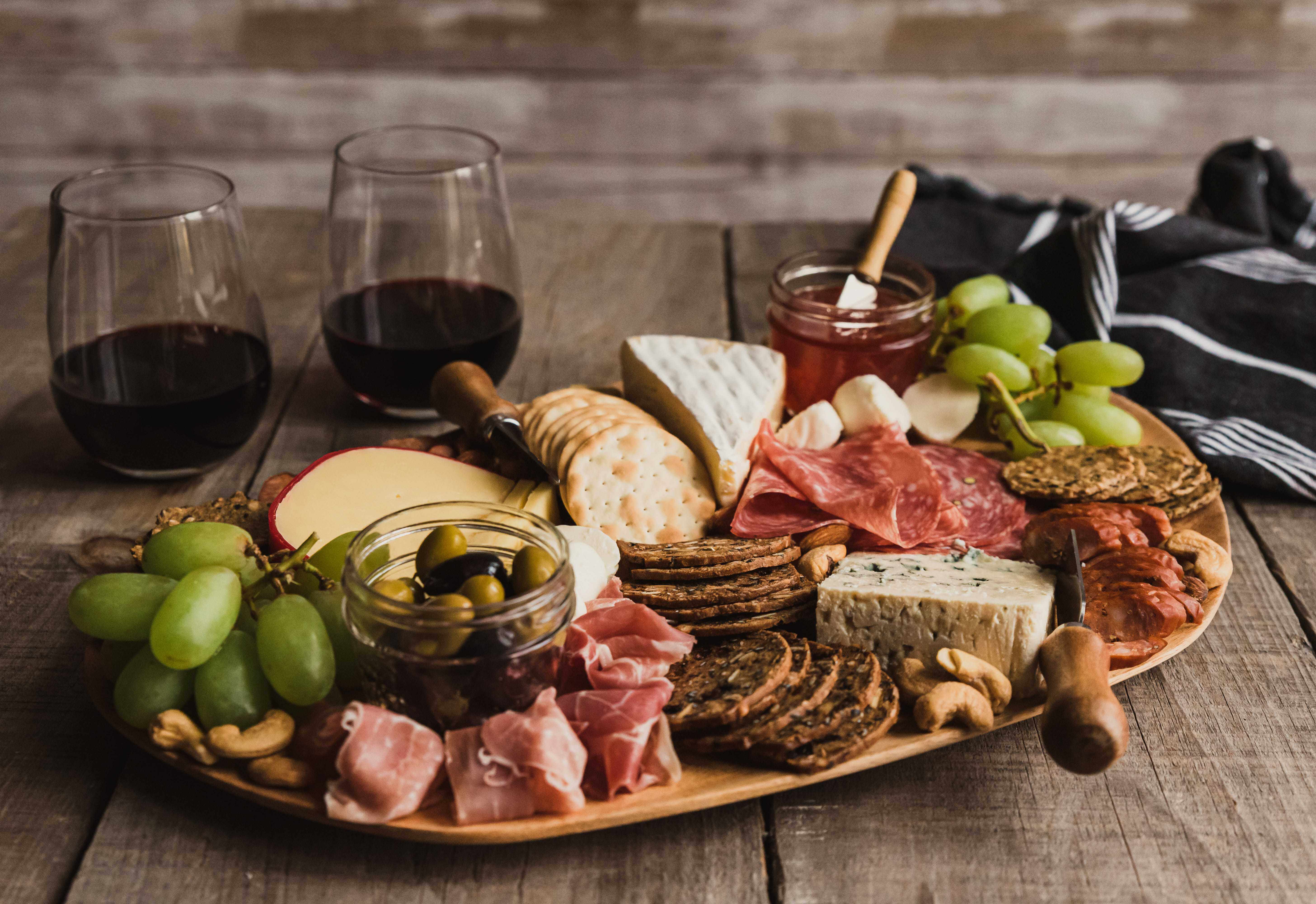 A wooden board with assorted cheeses, crackers, cured meats, green grapes, olives, and nuts, with two glasses of red wine on a rustic wooden table. A small jar of jam and a dark cloth napkin are also visible.