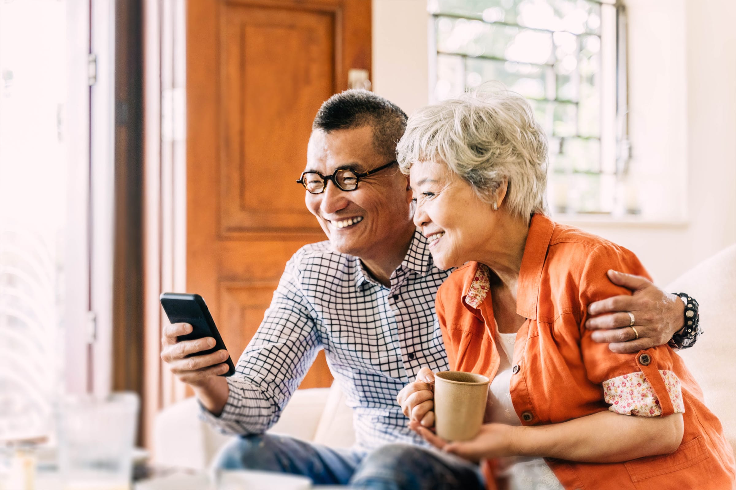 An older couple sits closely together on a couch, smiling as they look at a smartphone. The man has his arm around the woman, who is holding a mug. Sunlight streams in through a window behind them.