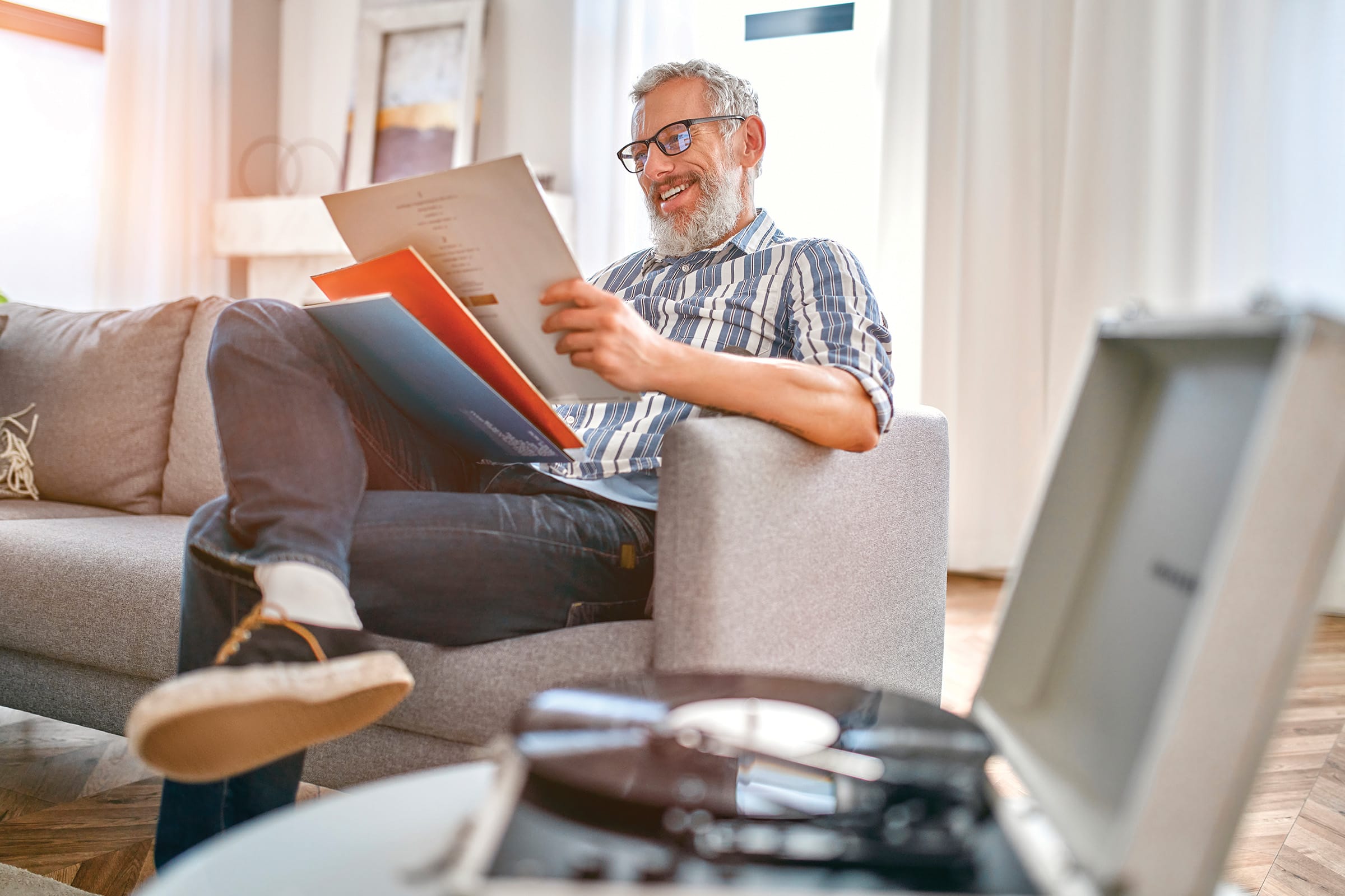 A smiling man with gray hair and a beard sits on a sofa, holding vinyl records and looking at one. A record player with a vinyl record is on a table in the foreground. The room is bright and cozy.