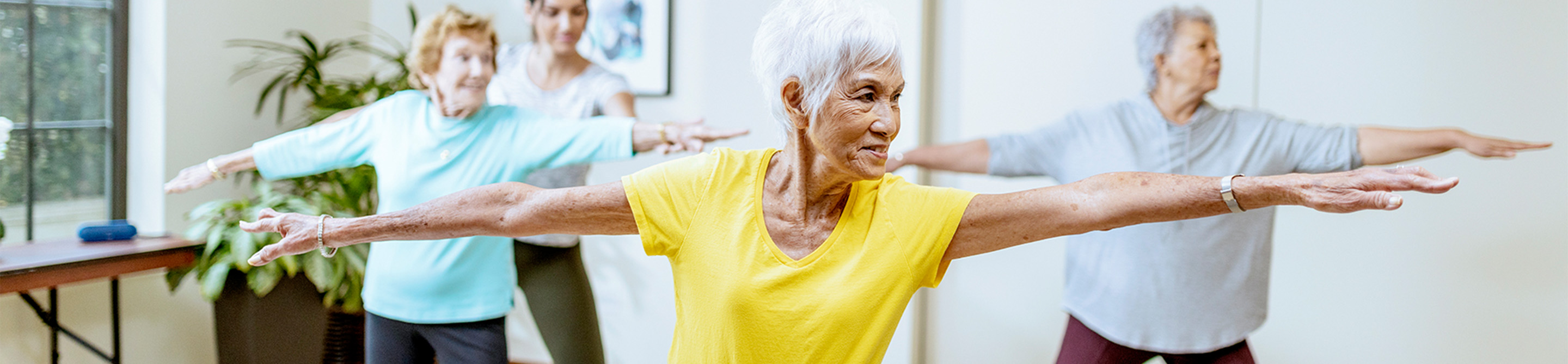 Three older women and an instructor participate in a group exercise class indoors, standing with arms extended, practicing balance and coordination. The woman in front wears a yellow shirt.