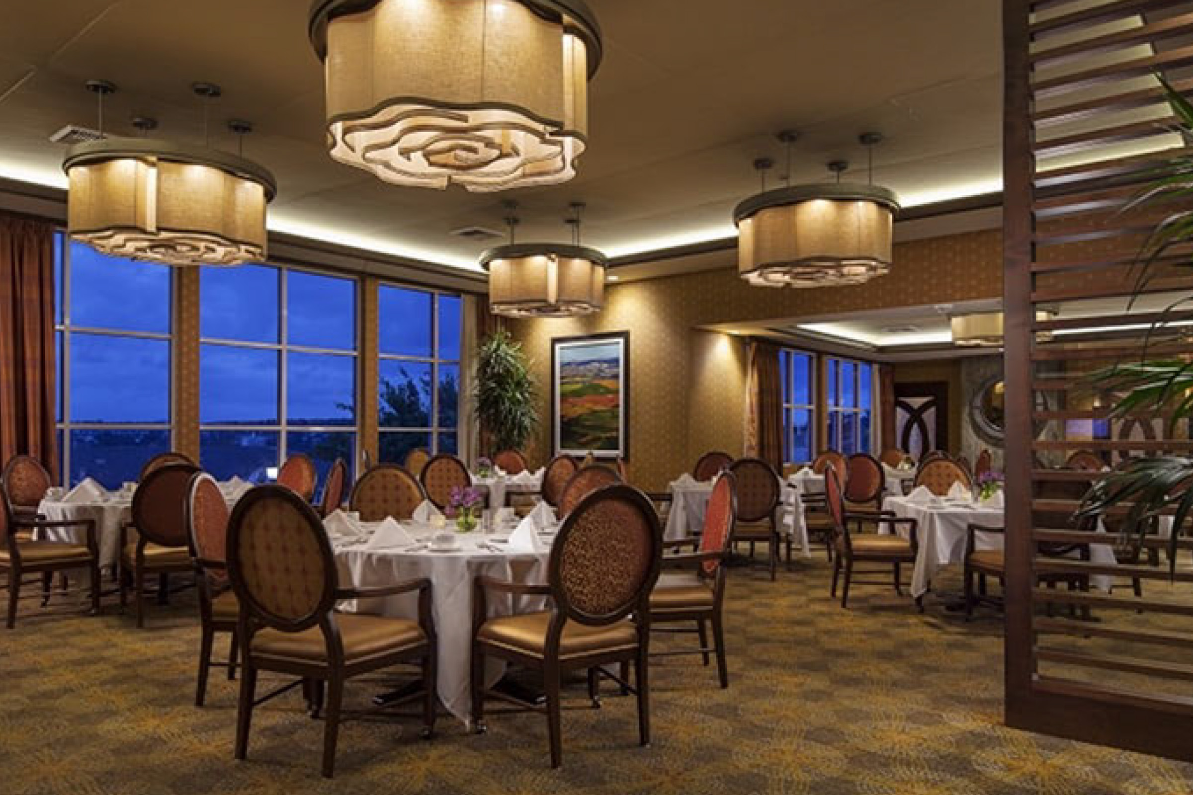 Elegant restaurant dining room with round tables set with white tablecloths and formal place settings. Large windows show a view of evening sky. Warm lighting and classic upholstered chairs create a cozy, upscale atmosphere.