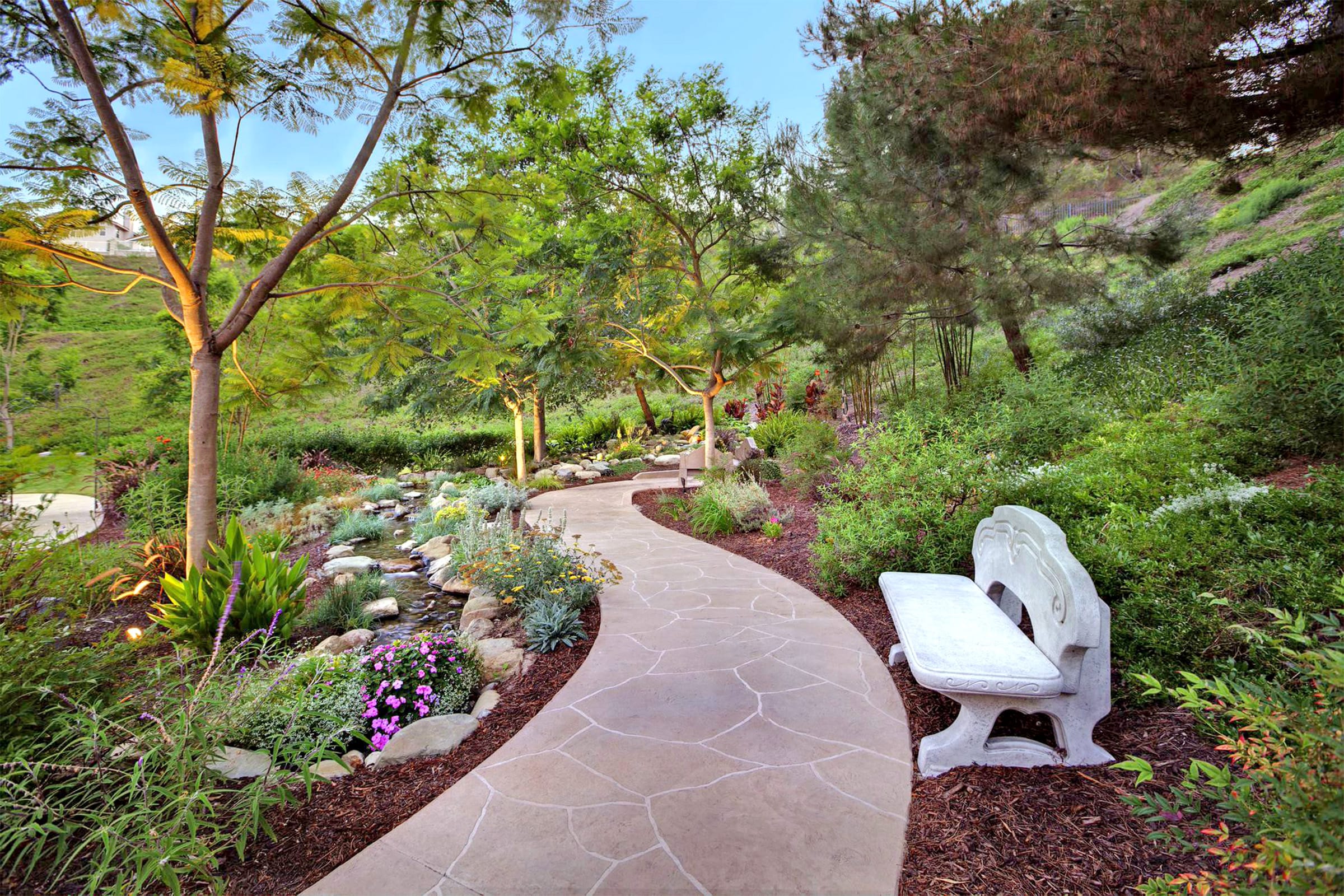 A winding stone path curves through a lush, green garden with flowering plants, tall trees, and a white stone bench on the right side. The scene is tranquil and inviting, with soft sunlight filtering through the trees.